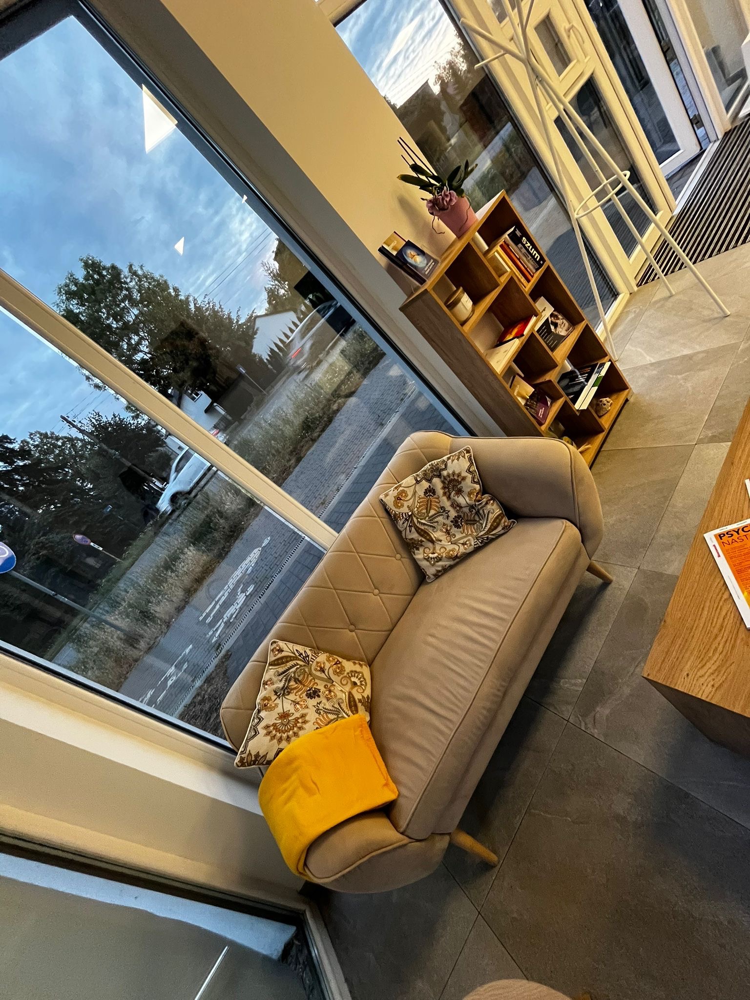 Jasne wnętrze poczekalni: beżowa sofa z poduszkami, regał z książkami, widok na ulicę przez duże okno, stolik z czasopismem.