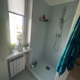 Marcin Zyga - Jasna łazienka po remoncie: kabina prysznicowa z odpływem liniowym, okno z roletą i dekoracjami. Błękitne ściany, biały grzejnik. Widok z góry.