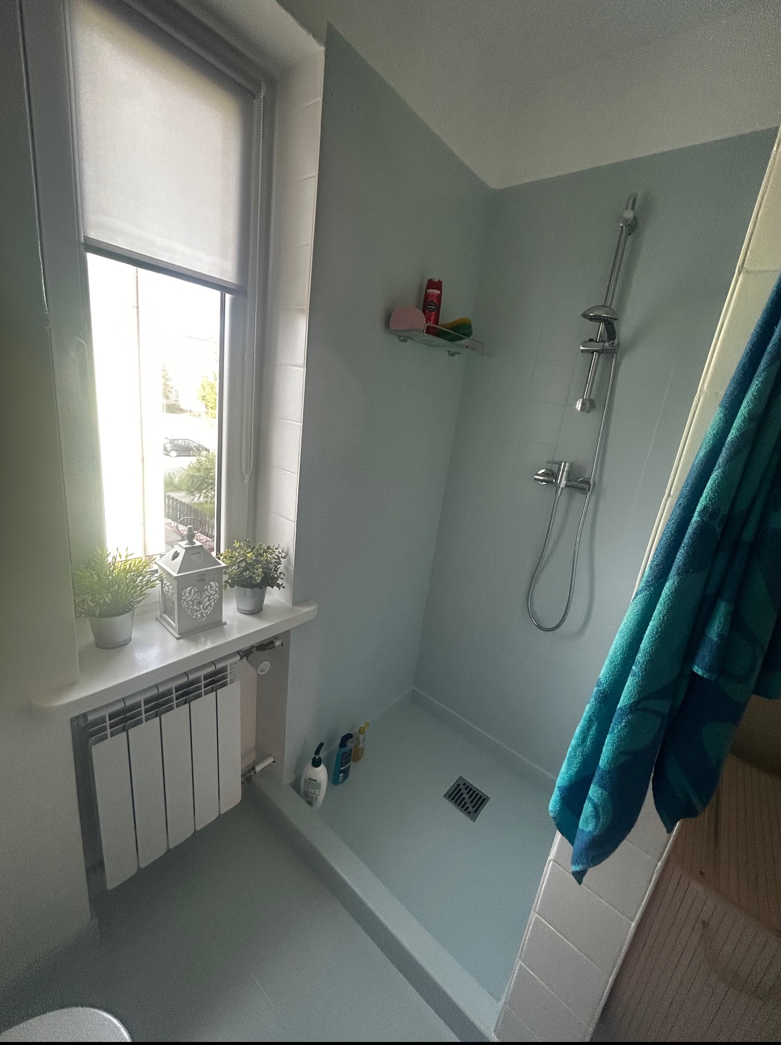 Jasna łazienka po remoncie: kabina prysznicowa z odpływem liniowym, okno z roletą i dekoracjami. Błękitne ściany, biały grzejnik. Widok z góry.