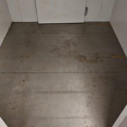 ABK-Reinigungsunternehmen - Brudna podłoga w windzie towarowej z metalowymi panelami i resztkami po budowie. Widoczne ślady po remoncie i żółte oznaczenia na ścianach.