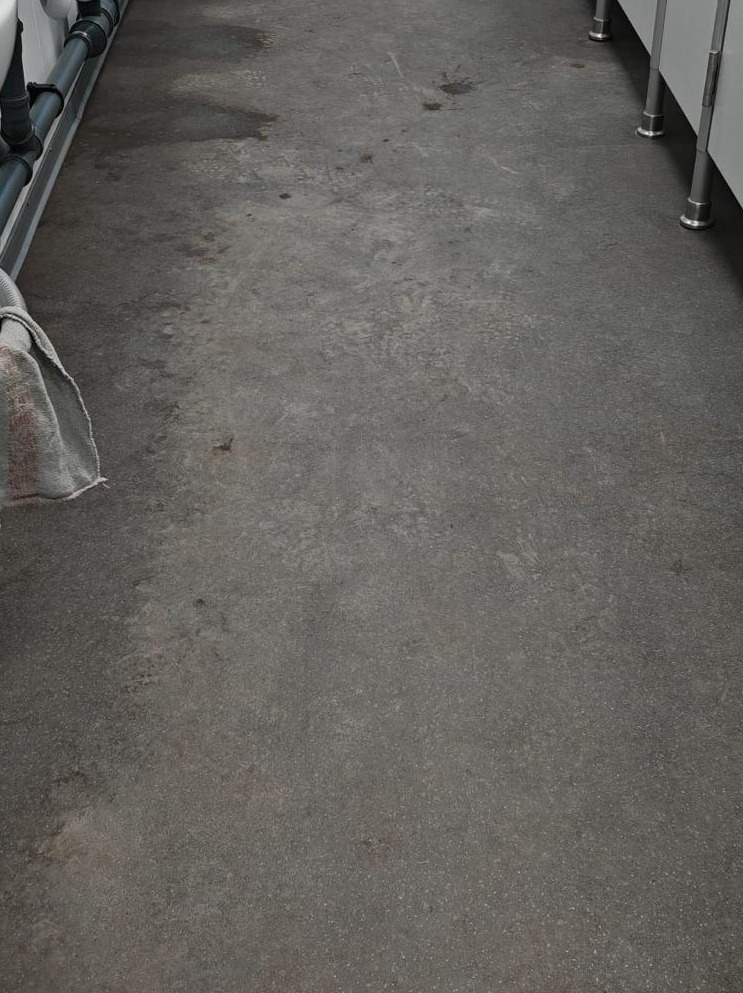 Szara, betonowa posadzka z plamami i zaciekami, widoczny fragment rury przy ścianie i fragment szafki. Posadzka wymaga czyszczenia.