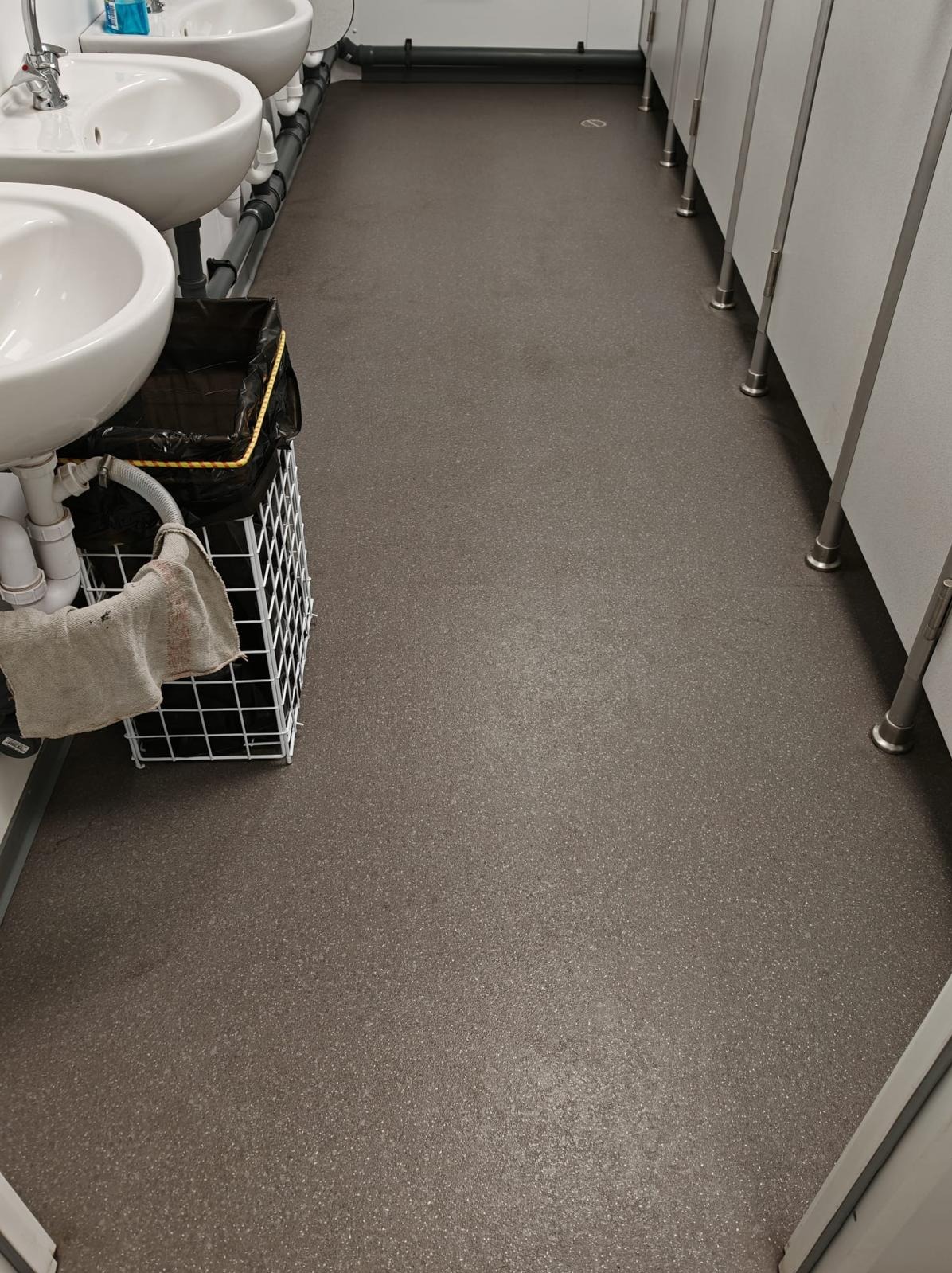 Czysta, szara podłoga w toalecie publicznej z umywalkami i kabinami. Widoczny kosz na śmieci i szmata. Ujęcie z góry, ukazujące porządek i higienę.