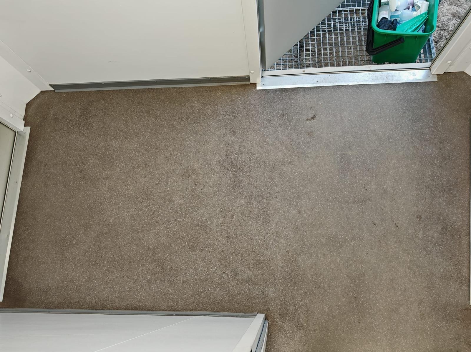 Wnętrze po sprzątaniu: gładka, brązowa podłoga, białe ściany i fragment zielonego wiadra z akcesoriami do sprzątania w rogu. Widok z góry.