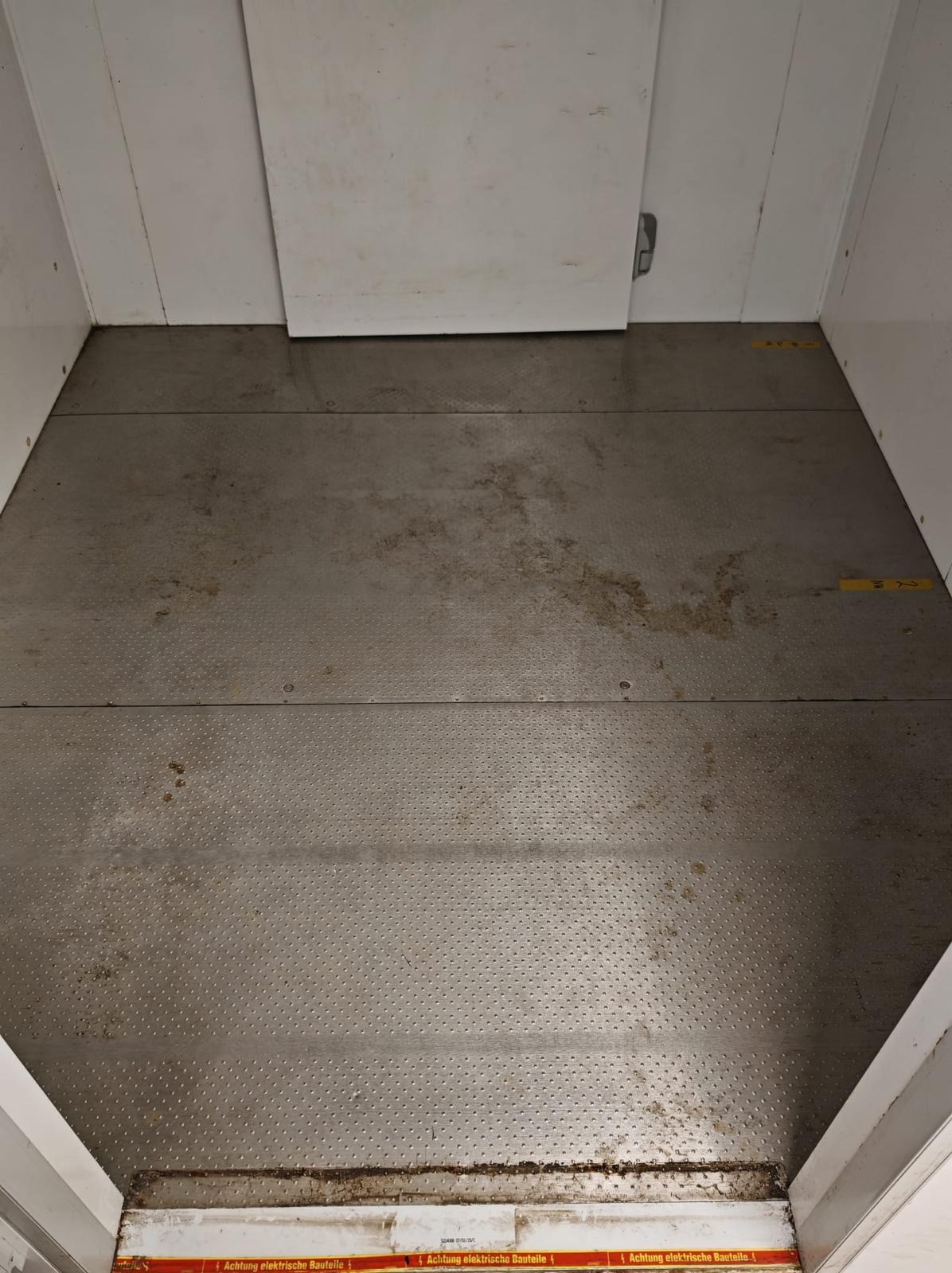 Brudna podłoga w windzie towarowej z metalowymi panelami i resztkami po budowie. Widoczne ślady po remoncie i żółte oznaczenia na ścianach.