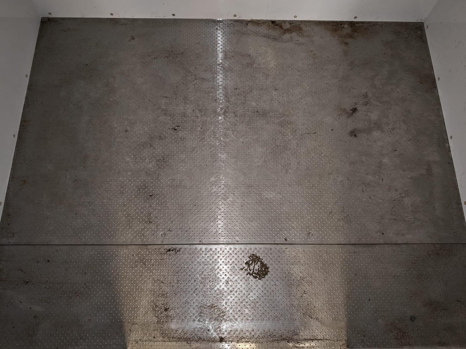 Metalowa podłoga z tłoczeniami, widoczne zabrudzenia i ślady użytkowania. Dwa poziomy podłogi, górny bardziej zabrudzony. Białe ściany po bokach.