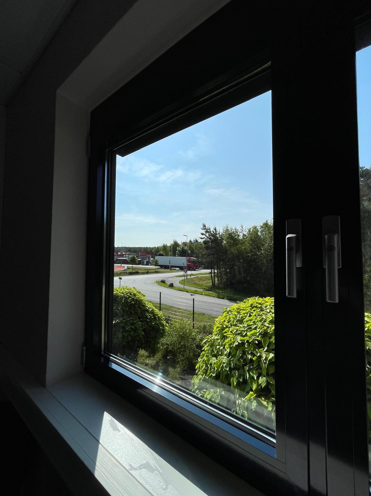 Ciemna rama okienna z widokiem na zieloną roślinność, drogę i błękitne niebo. Dwa srebrne uchwyty okienne po prawej stronie. Słoneczny dzień.