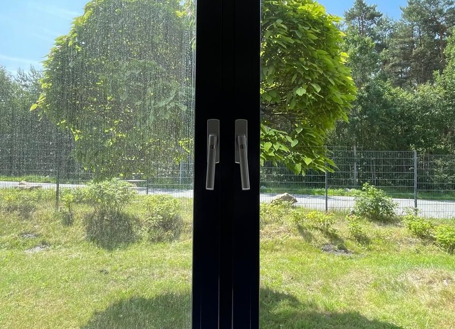 Częściowo umyte okno z widokiem na zieleń. Po lewej stronie szyba jest brudna, po prawej czysta. Widoczne klamki okienne i rama okienna.