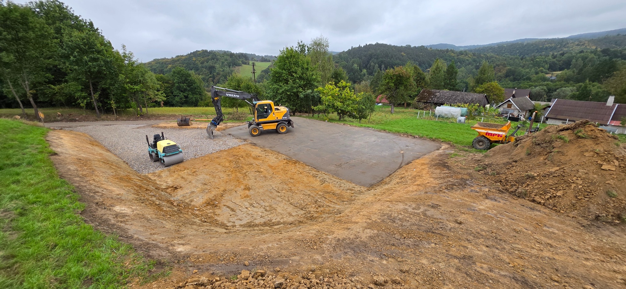 Żółty walec i koparka Volvo na wyrównanym terenie z widokiem na pagórkowaty krajobraz. Częściowo utwardzona powierzchnia, maszyny budowlane w akcji.