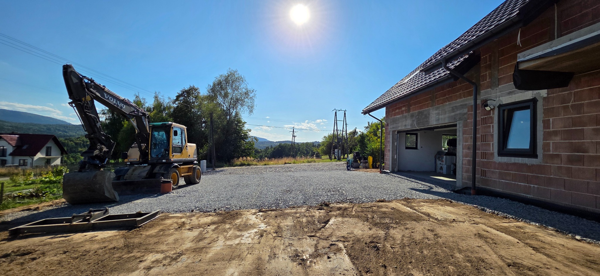 Żółta koparka Volvo na żwirowym podłożu przed budynkiem w trakcie budowy, w tle zielone wzgórza i błękitne niebo z mocnym słońcem. Widoczny etap utwardzania terenu.