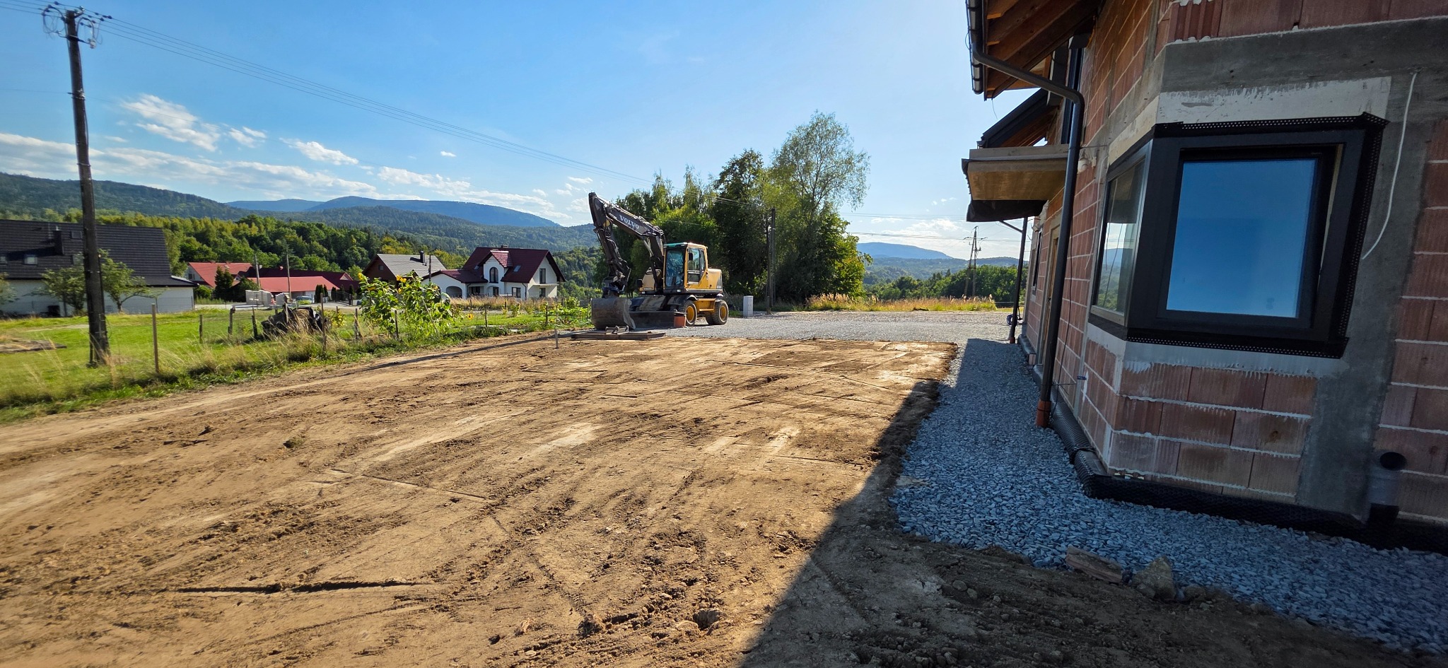 Żółta koparka Volvo na wyrównanym terenie budowy obok domu w stanie surowym otwartym, w tle góry i błękitne niebo.