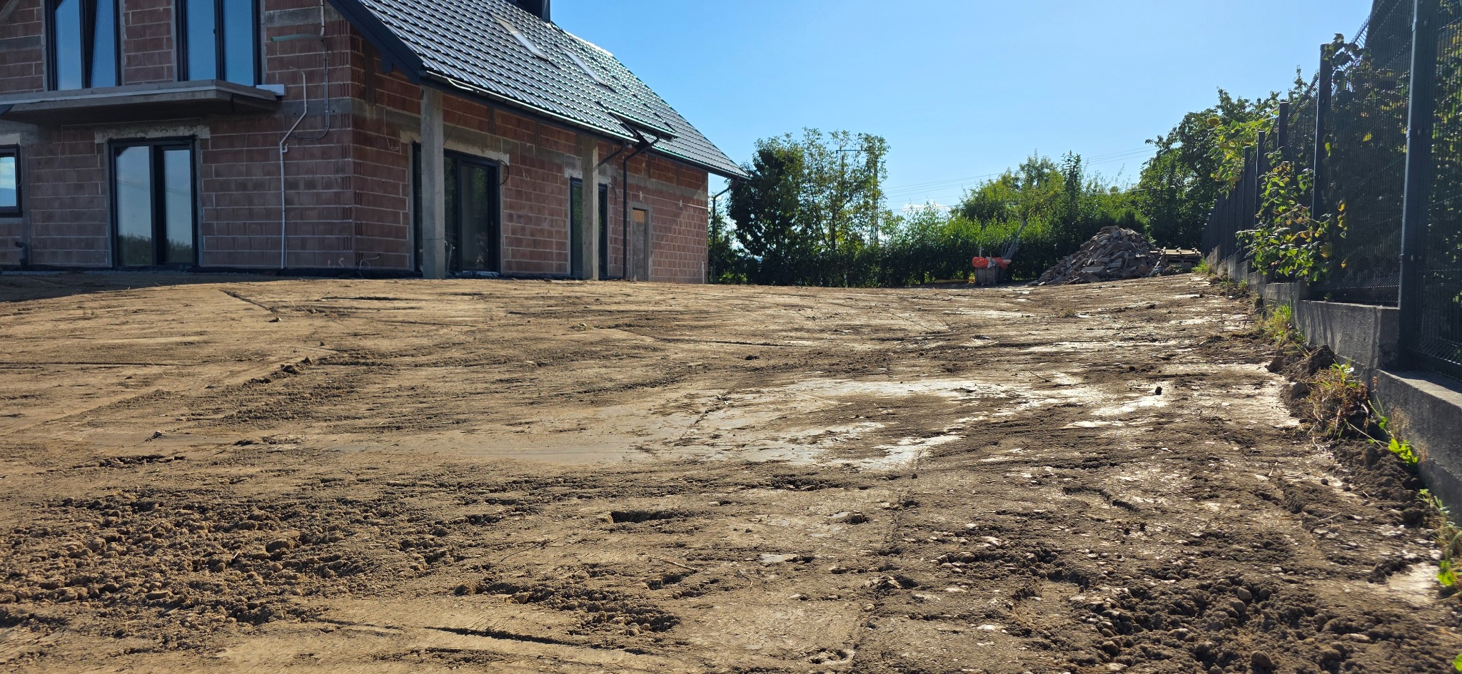 Nierówny, wyrównany teren pod budowę domu jednorodzinnego w stanie surowym otwartym. Widoczne ślady opon i hałda ziemi przy ogrodzeniu.