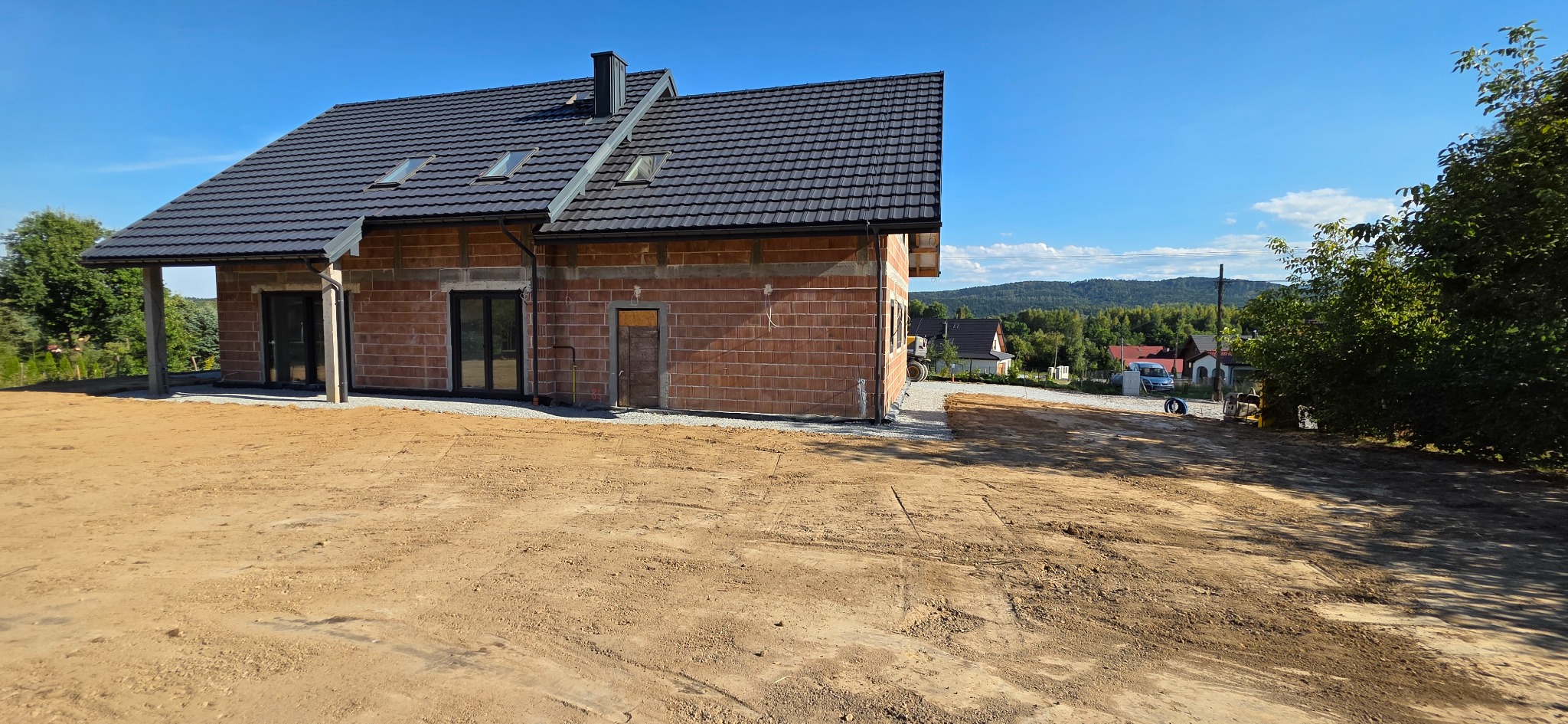Surowy stan domu z cegły z ciemnym dachem na wyrównanym terenie, widok na wzgórza w tle, słoneczny dzień.