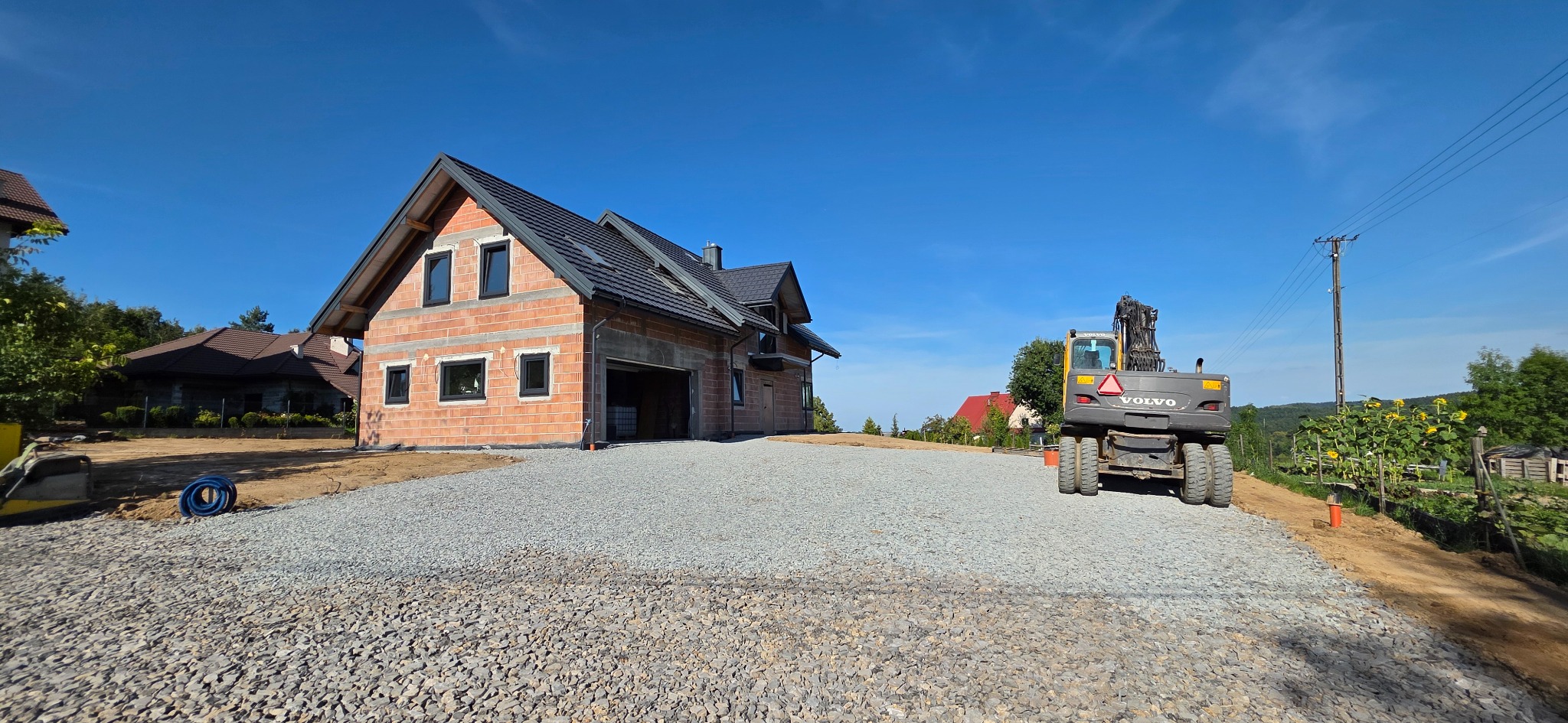 Nowo wybudowany dom z czerwonej cegły z utwardzonym żwirowym podjazdem i koparką VOLVO w tle, w słoneczny dzień.