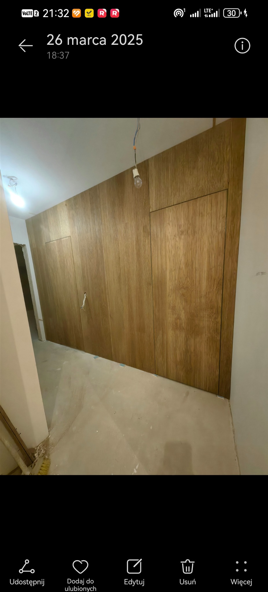 Ściana wykończona panelami drewnianymi z ukrytymi drzwiami, widoczne kable od oświetlenia, surowy beton na podłodze. Detal wnętrza w trakcie wykańczania.