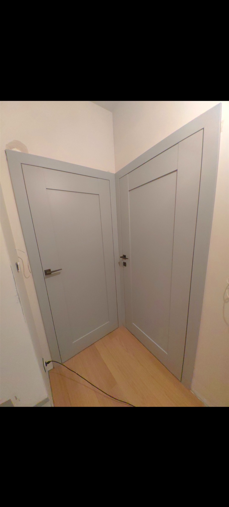 Dwa szare, wewnętrzne drzwi z prostokątnymi panelami, zamontowane pod kątem prostym, na drewnianej podłodze. Widoczny fragment ściany i gniazdko elektryczne.