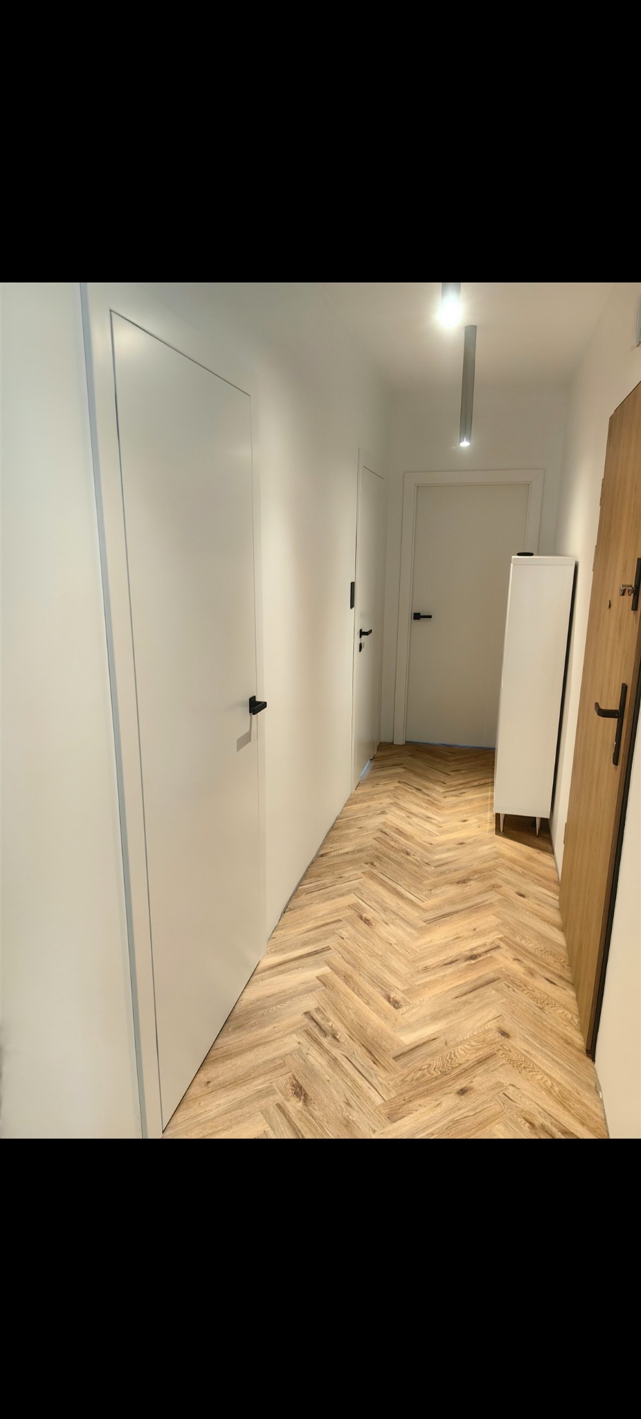 Korytarz z parkietem ułożonym w jodełkę. Widoczne białe drzwi z czarnymi klamkami. Oświetlenie sufitowe. Częściowo widoczna drewniana ościeżnica.
