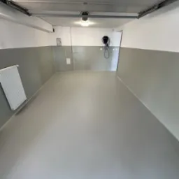 Szara posadzka żywiczna w garażu z instalacją do ładowania aut elektrycznych. Gładka powierzchnia i odpływ liniowy przy bramie garażowej.