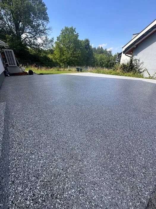 Szara, błyszcząca posadzka żywiczna na zewnątrz budynku w słoneczny dzień, z widoczną fakturą i elementami otoczenia.