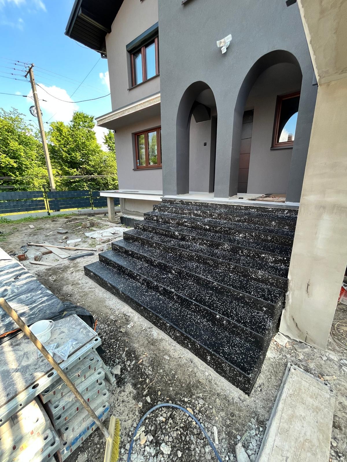 Nowoczesne, czarne schody wejściowe z białymi akcentami, prowadzące do szarego domu z łukowym wejściem. Widoczne materiały budowlane.