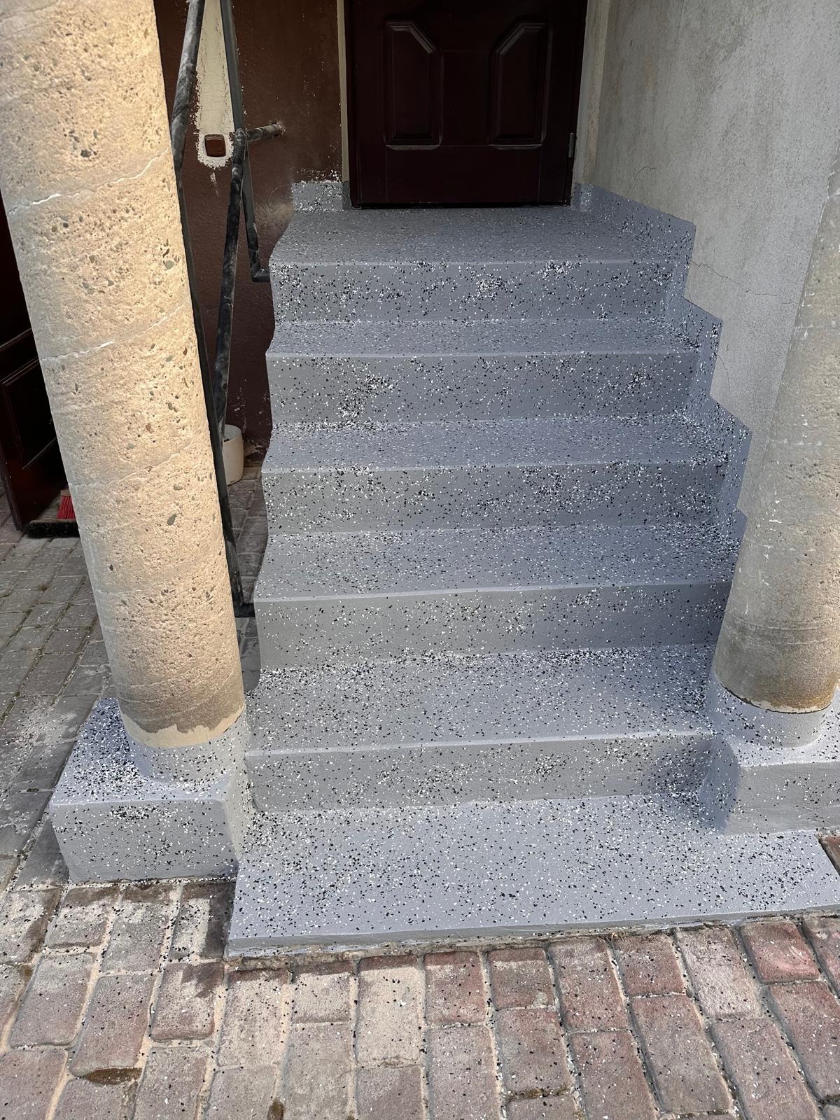 Schody wejściowe z szarą posadzką żywiczną z kolorowymi drobinkami, prowadzące do brązowych drzwi, otoczone kolumnami. Widok na całe schody.