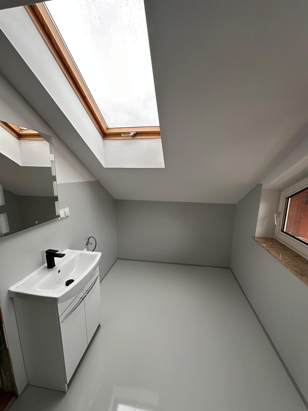 Jasnoszara łazienka na poddaszu z oknem dachowym, nową białą szafką z umywalką i czarną armaturą. Świeżo pomalowane ściany i podłoga z połyskiem.