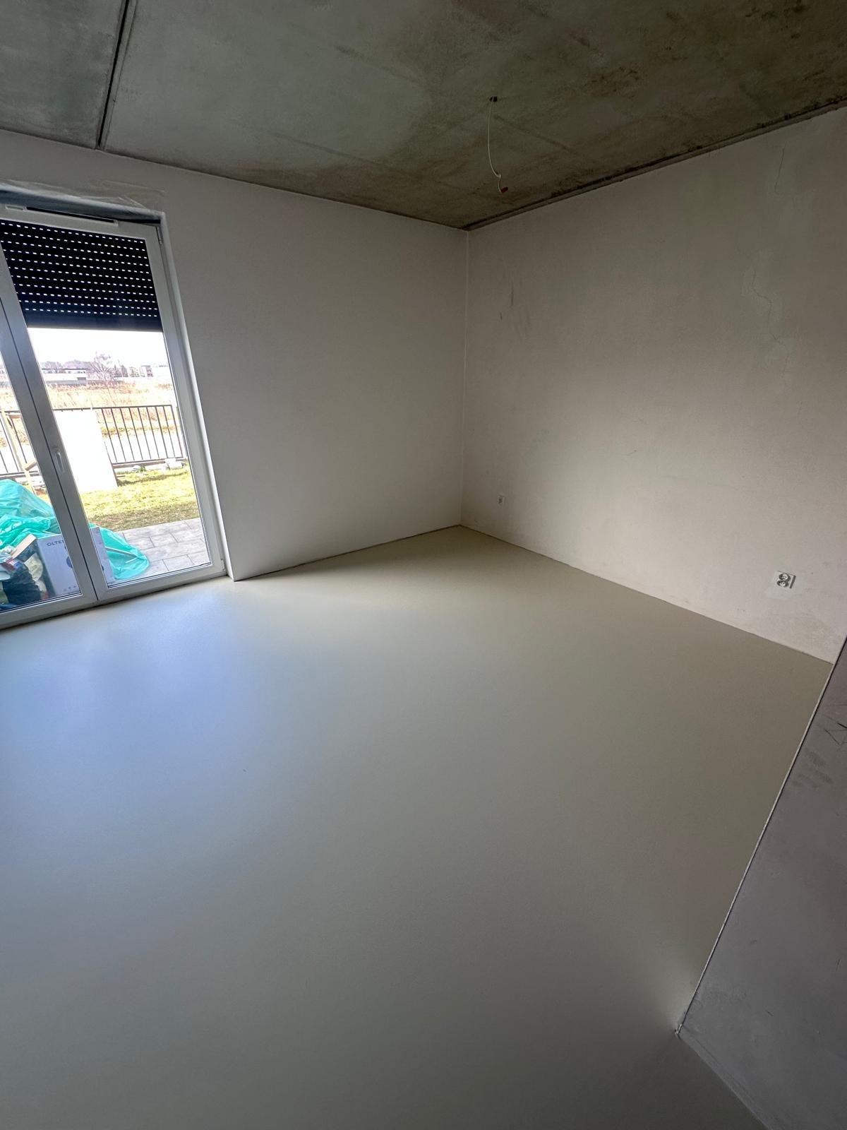 Jasne, gładkie posadzki żywiczne w surowym wnętrzu z betonowym sufitem i oknem z roletami. Widoczne puszki elektryczne na ścianie. Minimalistyczny design.