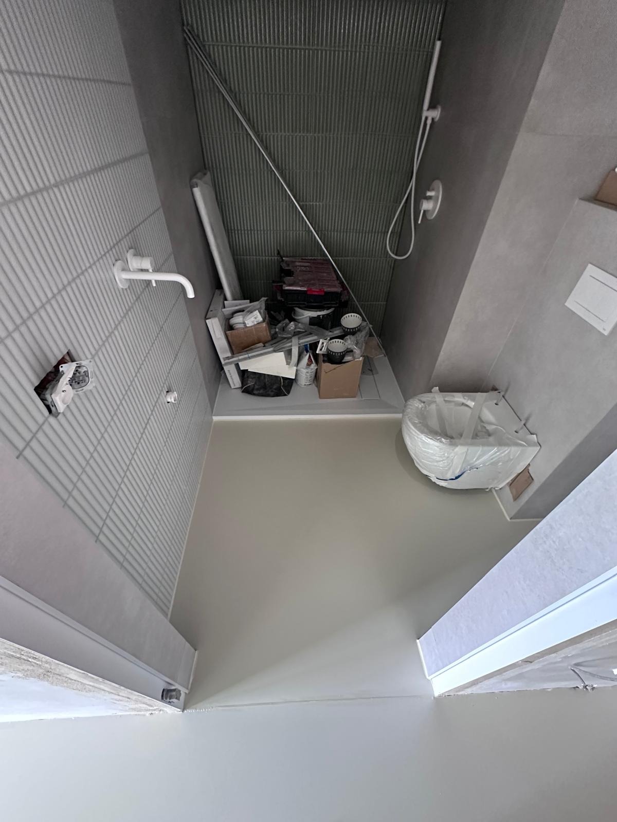 Wnętrze łazienki w trakcie remontu: położona jasna posadzka, częściowo wykończone ściany z szarymi płytkami, armatura i materiały budowlane. Widok z góry.