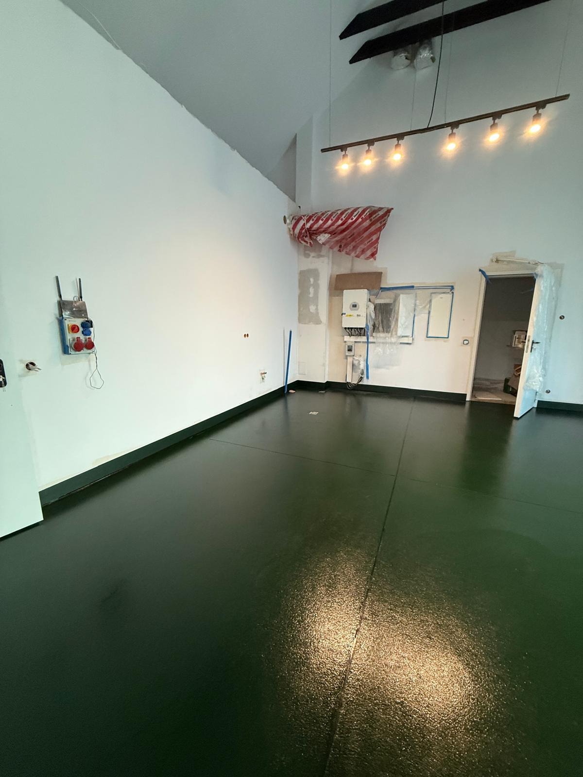 Błyszcząca, ciemnozielona posadzka w przestronnym, jasnym pomieszczeniu z białymi ścianami. Widoczne zabezpieczenia folią malarską i instalacje elektryczne.