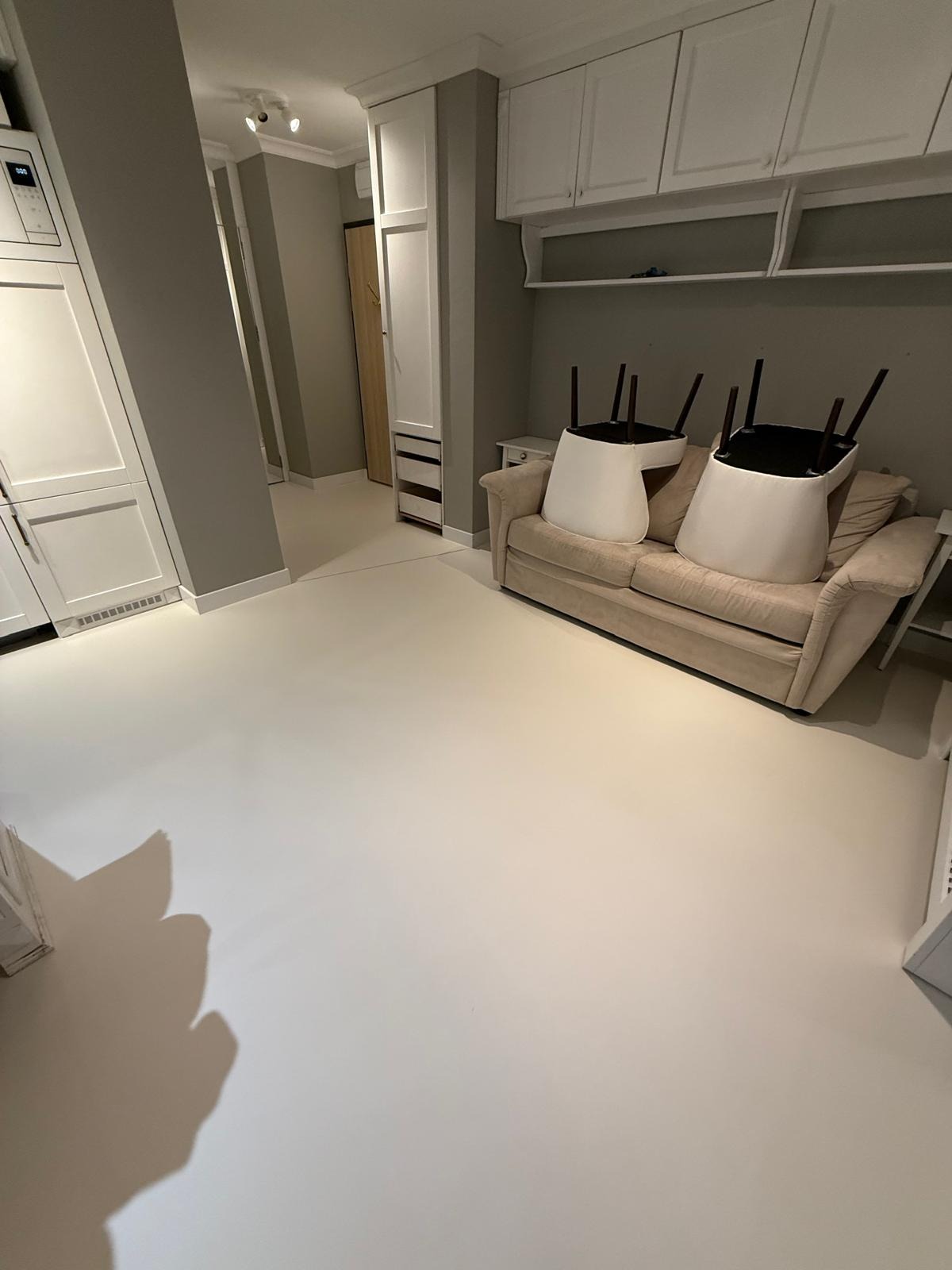 Jasna, gładka posadzka w pokoju dziennym z beżową sofą i odwróconymi krzesłami. Widoczne białe szafki i drzwi. Minimalistyczne wnętrze po remoncie.