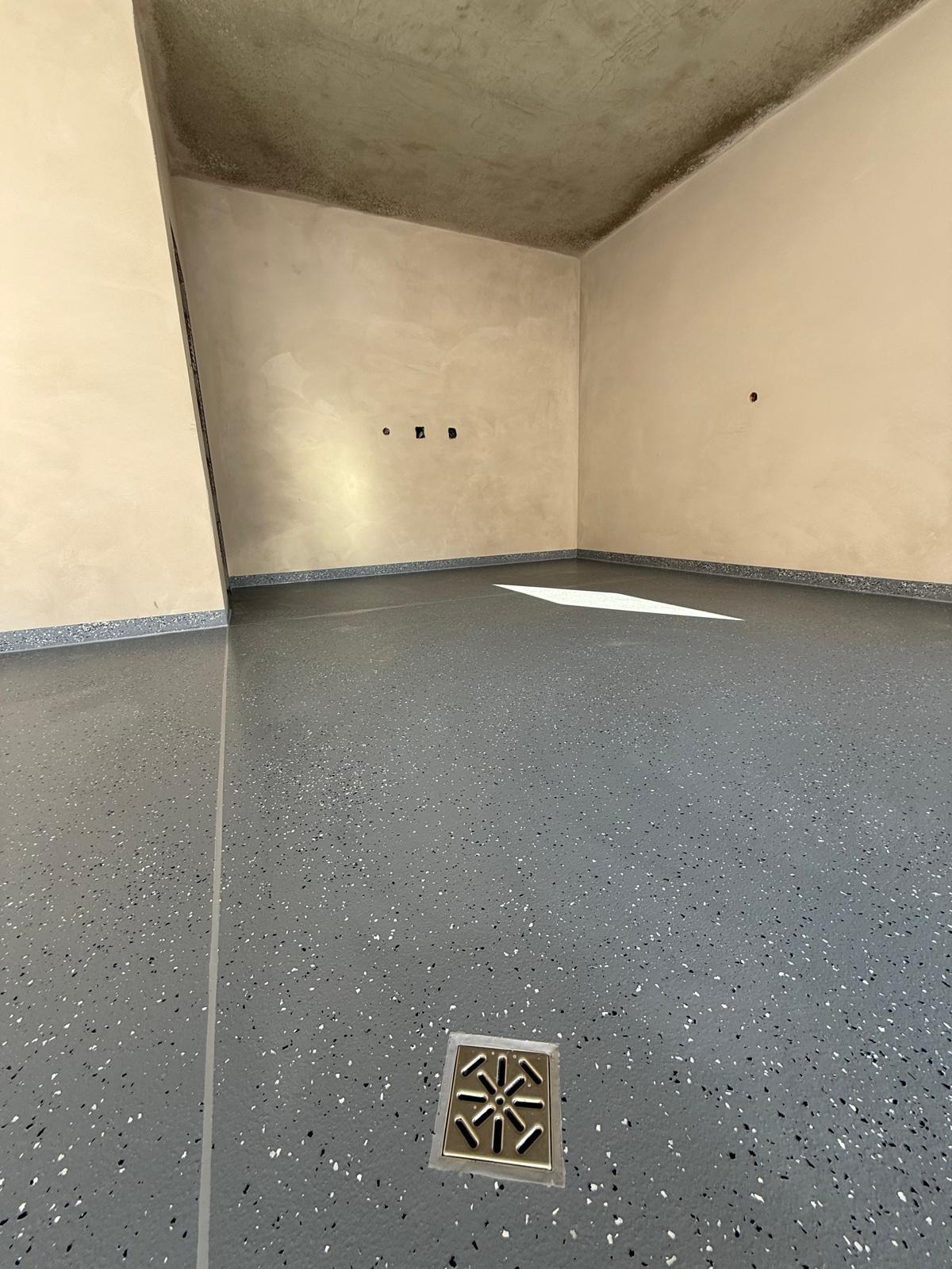 Szara posadzka żywiczna z brokatem w pomieszczeniu z beżowymi ścianami i odpływem podłogowym. Widoczne niedoskonałości ścian i sufitu.