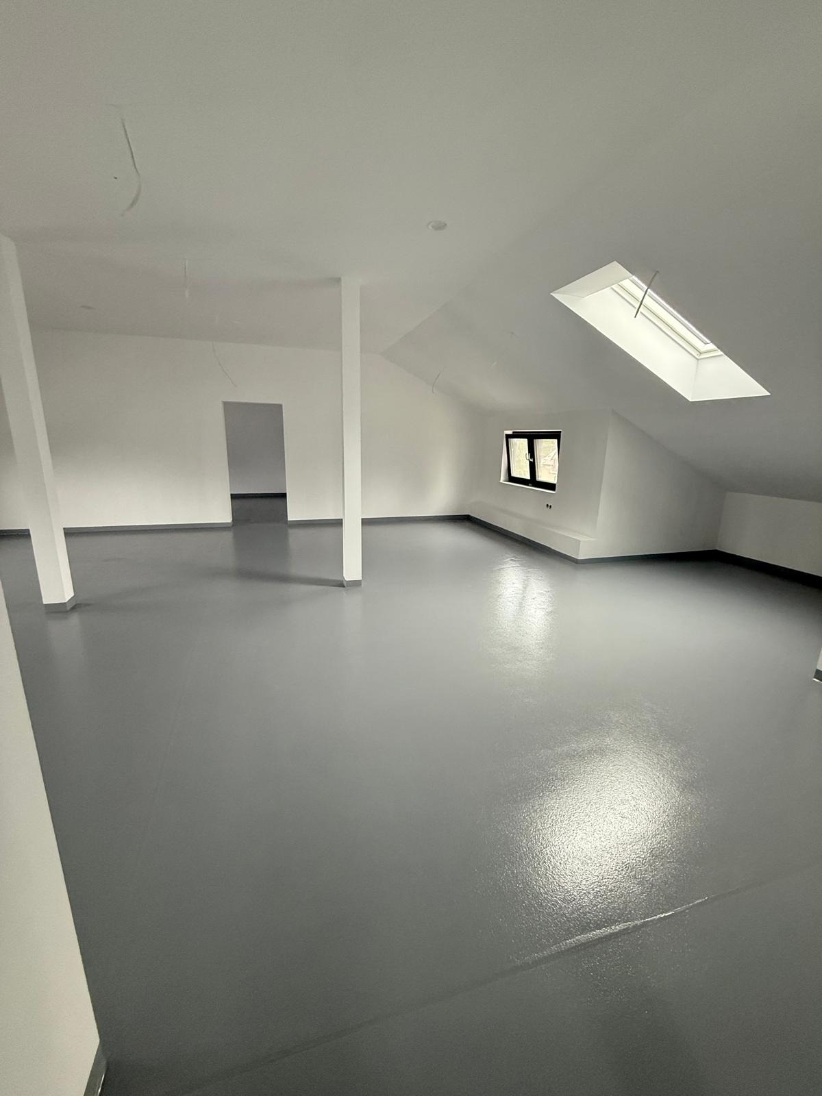 Szara, błyszcząca posadzka w jasnym, przestronnym pokoju na poddaszu z oknem dachowym i filarami. Minimalistyczne wnętrze po remoncie, świeżo wykończone.