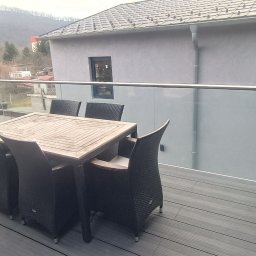 W-Grand Swietłana Makarewicz - Nowoczesny taras z deską kompozytową w odcieniach szarości, szklana balustrada, stół z krzesłami. W tle widok na góry i zabudowania.