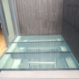 W-Grand Swietłana Makarewicz - Szklana podłoga w nowoczesnym wnętrzu, widok z góry. Czarne, drewniane ściany okalają konstrukcję. W tle widoczny fragment wnętrza z krzesłem.