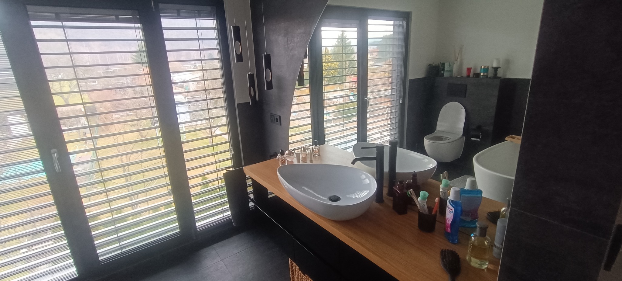 Nowoczesna łazienka z drewnianym blatem, owalnymi umywalkami i dużym oknem z żaluzjami, widok na zieleń za oknem. Minimalistyczne oświetlenie.