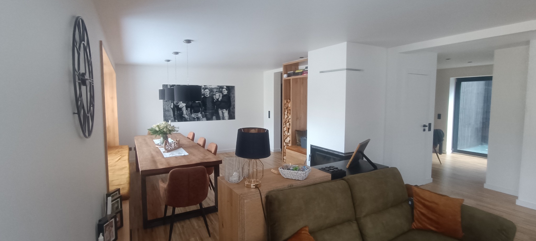 Jasne wnętrze z drewnianym stołem, krzesłami i sofą. Na ścianie duży zegar i czarno-białe zdjęcie. Widoczny kominek i drewniane półki.