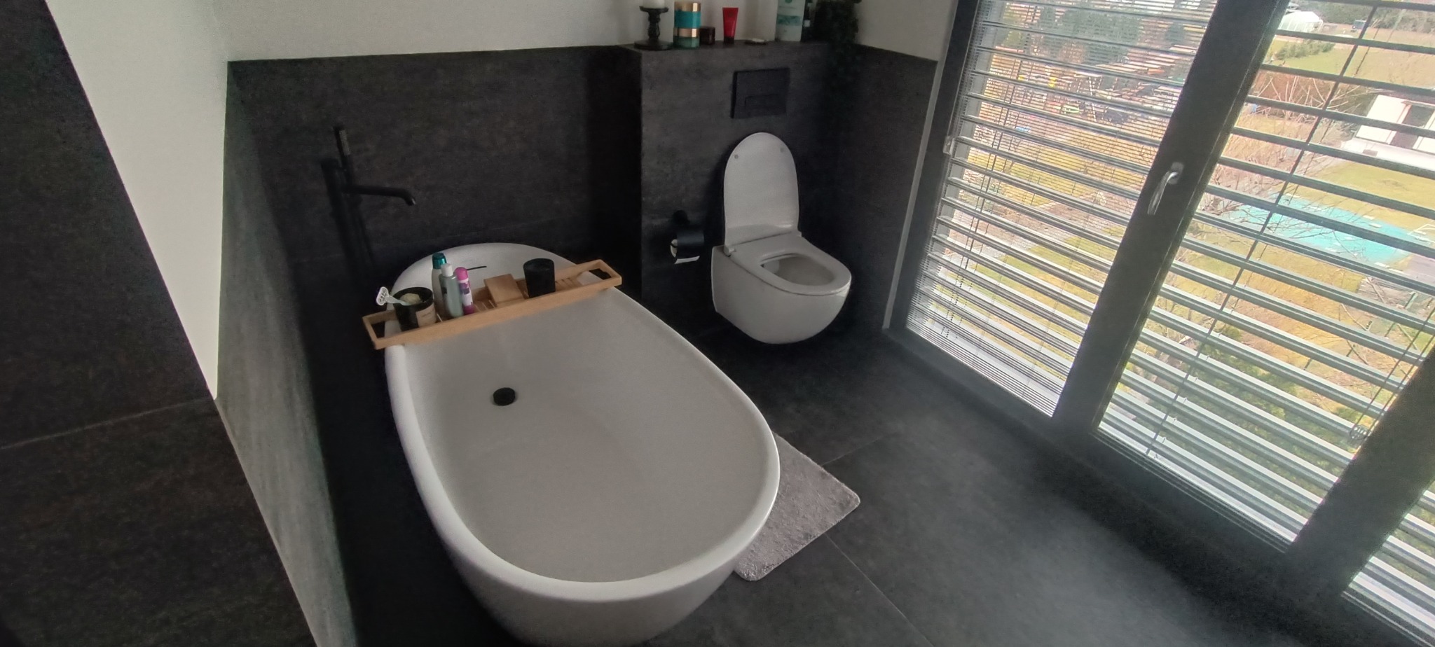 Nowoczesna łazienka z wolnostojącą wanną z akcesoriami, toaletą i dużym oknem z żaluzjami, utrzymana w ciemnej tonacji z elementami bieli.