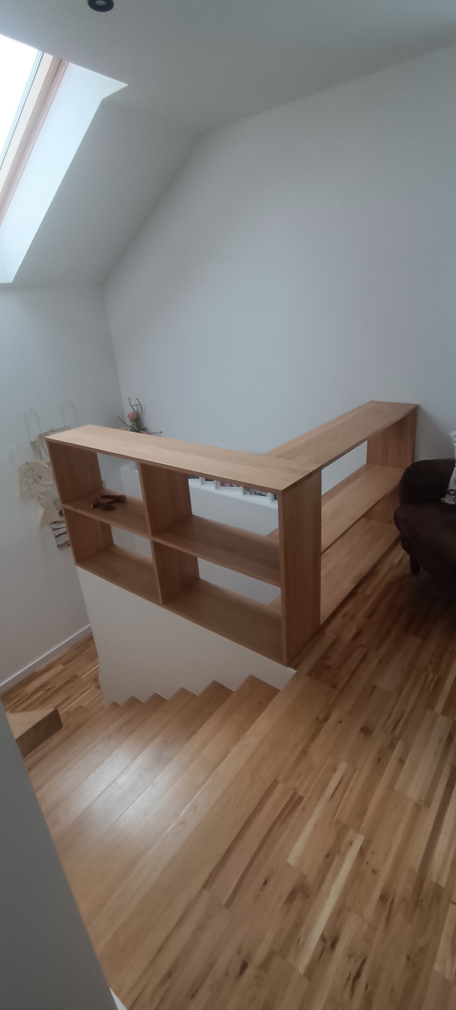 Drewniane schody z oryginalną balustradą, która pełni funkcję regału. Jasne drewno, minimalistyczny design, skosy poddasza.