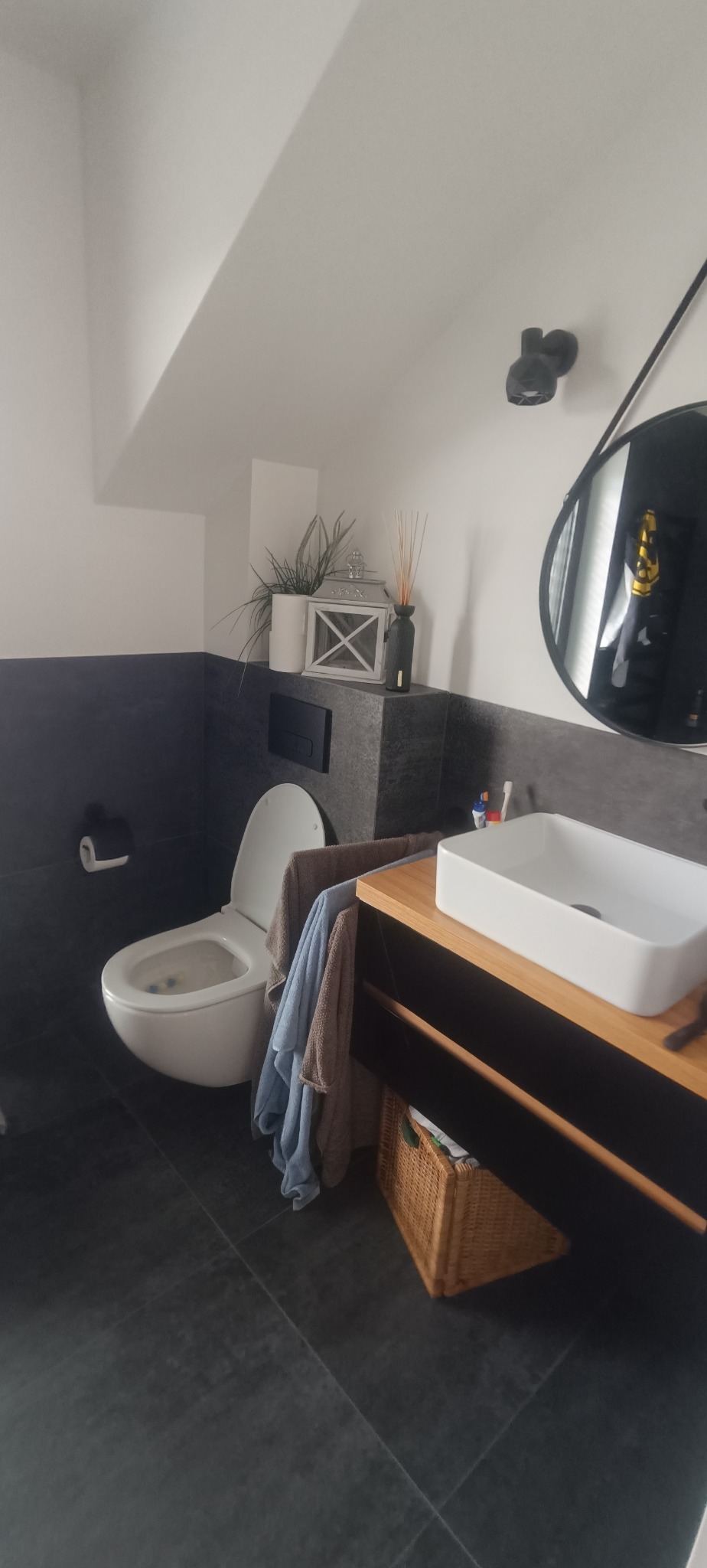 Nowoczesna łazienka w szarościach z drewnianymi akcentami. Umywalka nablatowa, okrągłe lustro i wisząca toaleta. Dekoracje: roślina, lampion i dyfuzor zapachowy.
