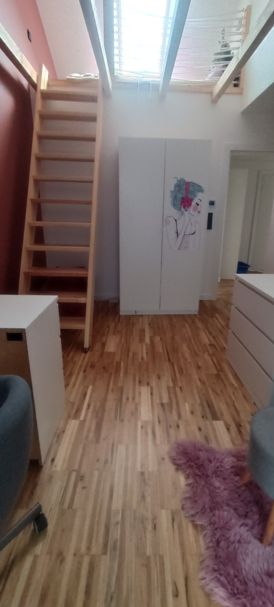 Jasne drewniane schody prowadzą do antresoli w pokoju. Biała szafa z rysunkiem dziewczyny w słuchawkach. Parkiet z naturalnego drewna.