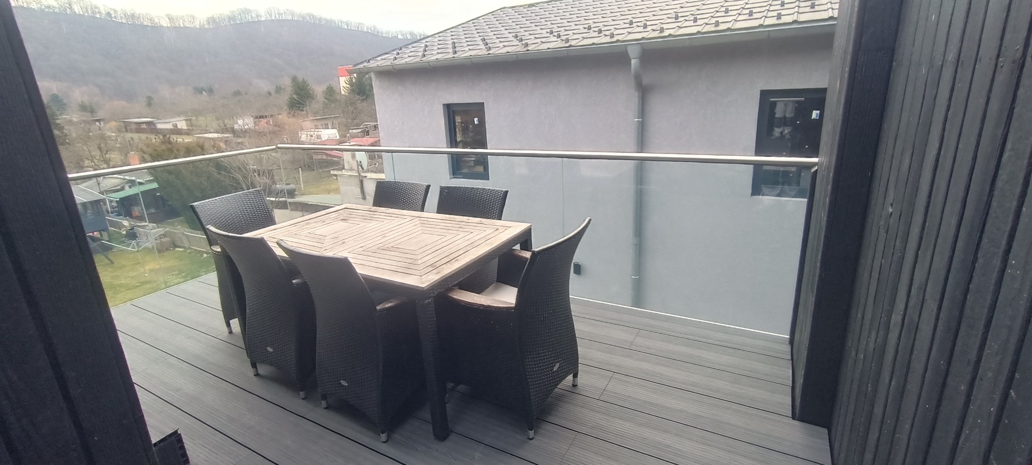Nowoczesny taras z deską kompozytową w odcieniach szarości, szklana balustrada, stół z krzesłami. W tle widok na góry i zabudowania.