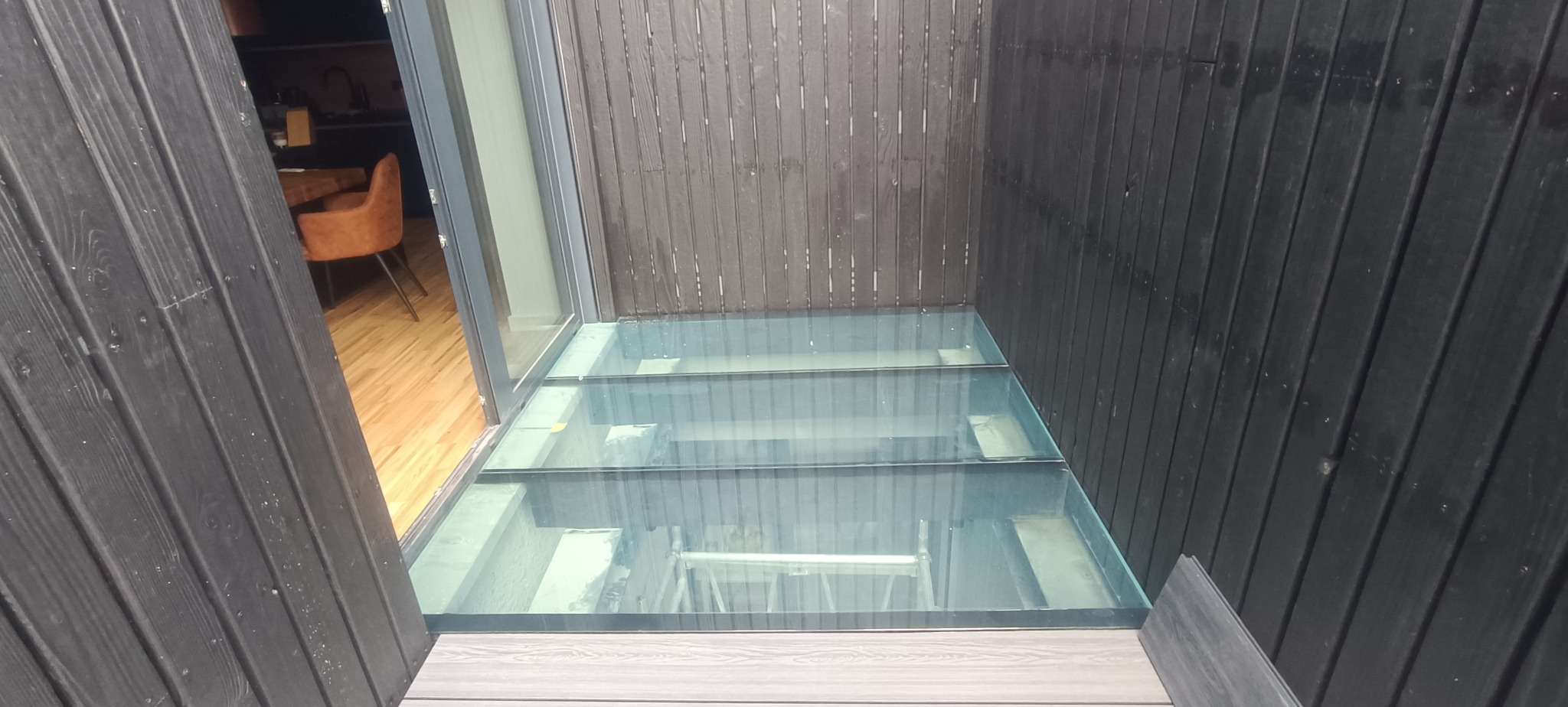 Szklana podłoga w nowoczesnym wnętrzu, widok z góry. Czarne, drewniane ściany okalają konstrukcję. W tle widoczny fragment wnętrza z krzesłem.
