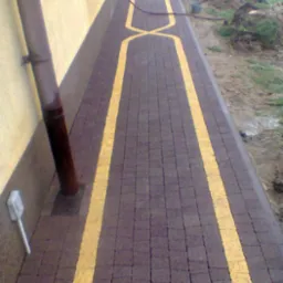 Ułożona, brązowa kostka brukowa z żółtymi pasami wzdłuż ścieżki i wzorem X w środku. W tle ziemia, trawa i maszyna budowlana. Widok z góry, perspektywa zbieżna.