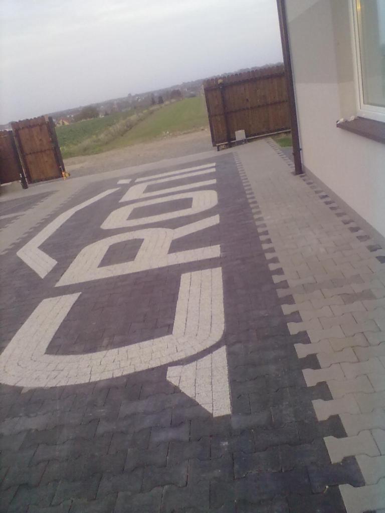 Ułożona kostka brukowa w ciemnych odcieniach z jasnym napisem. Widoczne otwarte, drewniane wrota i fragment budynku z oknem.