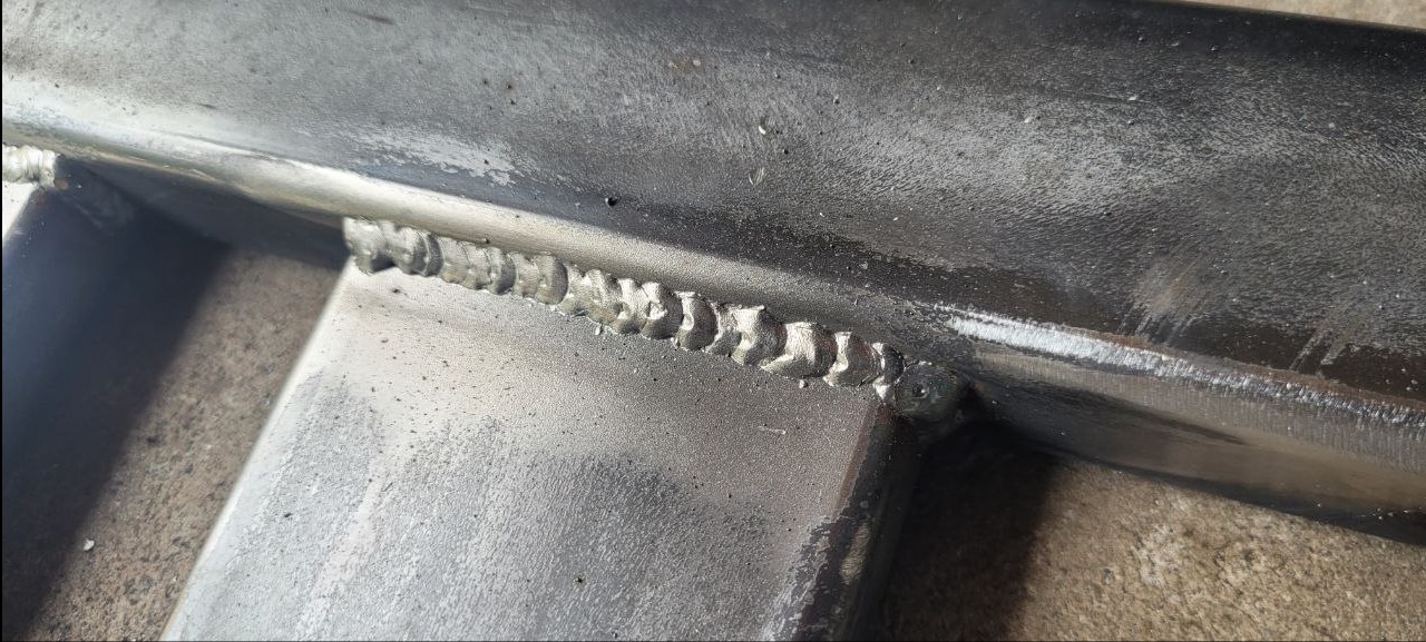 Szczegółowe zbliżenie spawu na metalowej konstrukcji. Widoczne regularne ślady spawania, srebrzysta powierzchnia metalu i szorstkie tło. Precyzyjne łączenie.