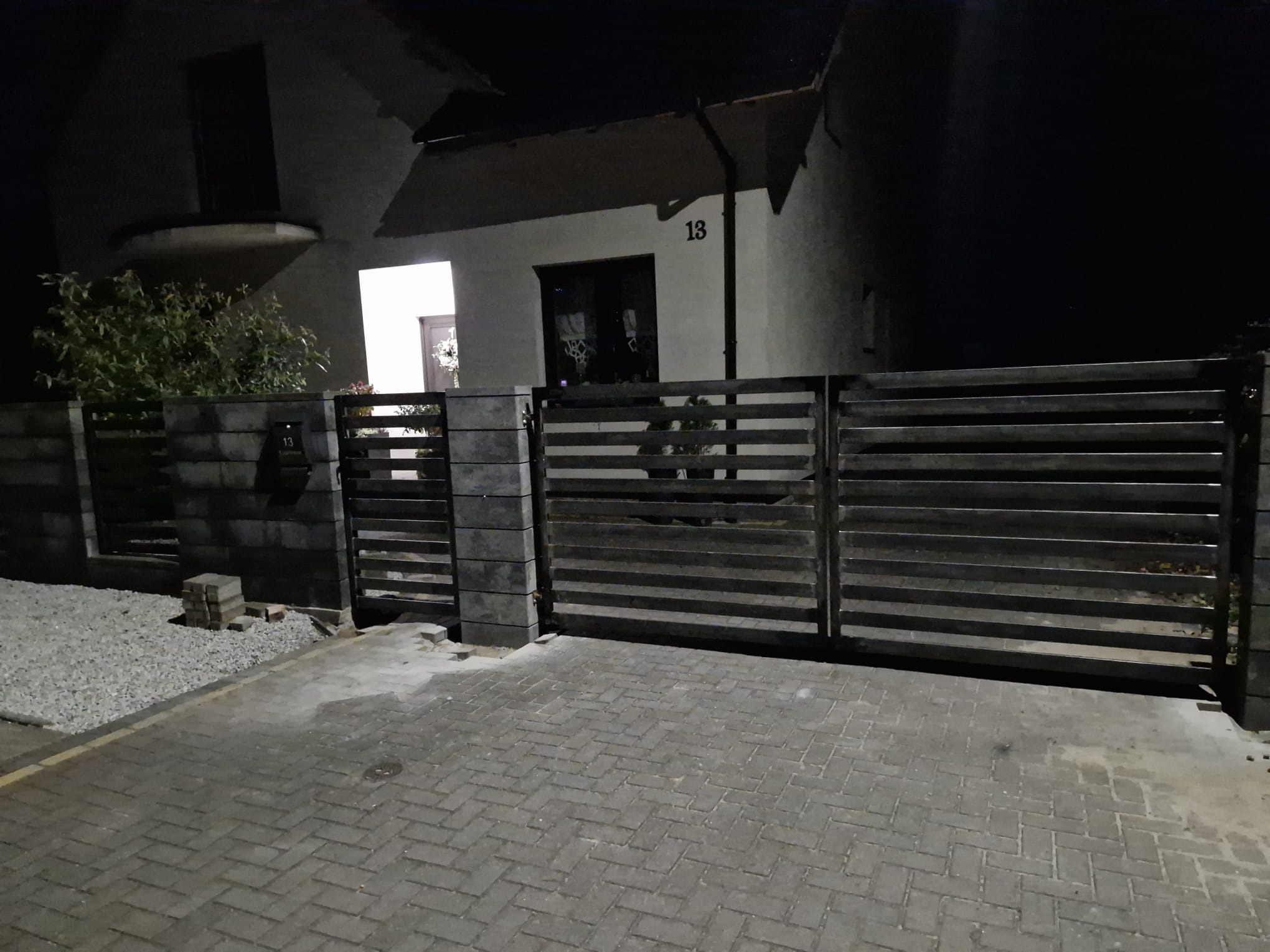Nowoczesna, pozioma brama wjazdowa z metalowymi przęsłami i betonowymi słupkami, oświetlona nocą, na tle domu z numerem 13.