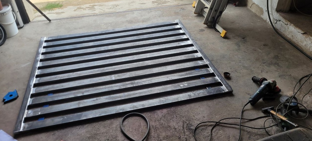 Metalowa konstrukcja kratowa z poziomych profili, leżąca na betonowej posadzce warsztatu. Widoczne narzędzia: szlifierka kątowa i młotek. Częściowo widoczna drabina.