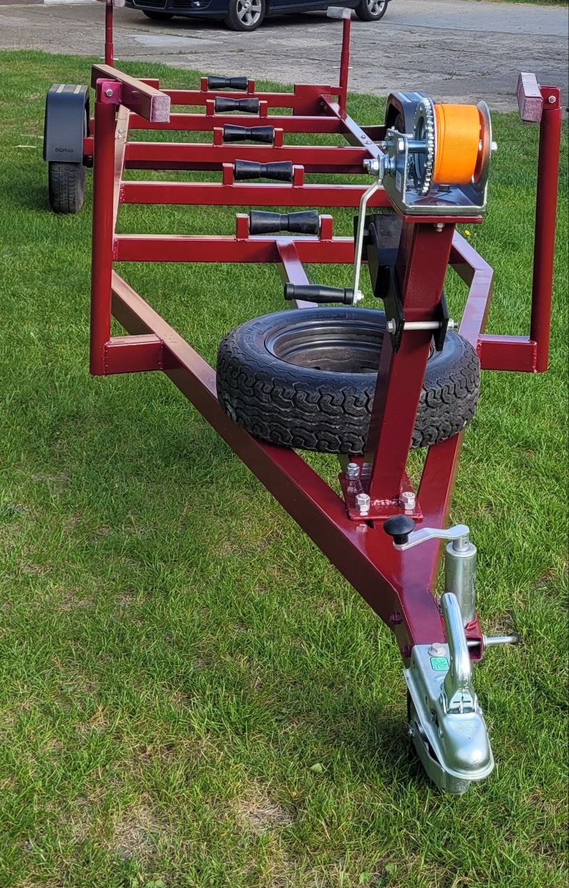 Czerwona, spawana przyczepa z kołem zapasowym i wciągarką, stojąca na trawie. Widoczny hak holowniczy i elementy konstrukcyjne. Detal wykonania.