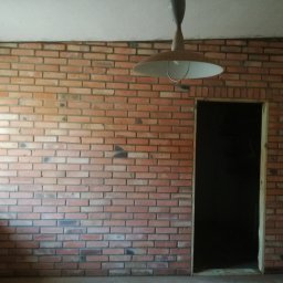 Mirbud - Ściana z cegły klinkierowej w pomieszczeniu z widocznym oknem i otworem drzwiowym, oświetlona wiszącą lampą. Wykończenie wnętrza w stylu industrialnym.