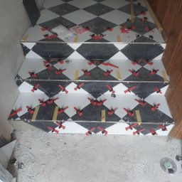 Mirbud - Układanie czarno-białych płytek w romby na schodach z użyciem czerwonych klipsów poziomujących. Widoczne fragmenty płytek i narzędzia.