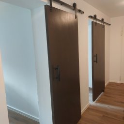 MIRBUD Mirosław Pastuszek - Dwa brązowe, przesuwne drzwi stodołowe z czarnymi uchwytami i szynami, zamontowane w białej ścianie. Widoczna podłoga z paneli i fragment drugiego pomieszczenia.